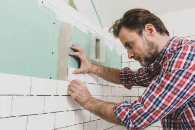 Kafkas iç mimar (tiler) duvarda küçük beyaz seramik fayanslar sergiliyor. Banyo tadilatı işe yarıyor. Ekose gömlekli tamirci..