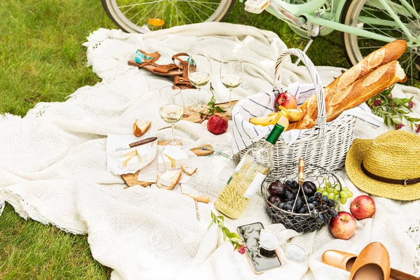 French style outdoor picnic on the white blanket. Wicker basket with baguettes, wine, fruits and camembert cheese. Pastel green bicycle in the background. Summer leisure - lifestyle.