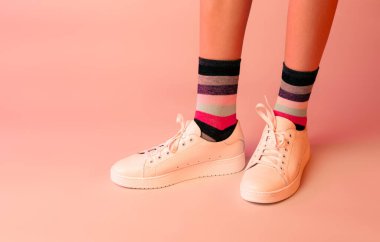 Moda - beyaz deri spor ayakkabılar, renkli çizgili çoraplar ve kız bacakları. Pastel pembe arka planda ayakkabılar. Boş kopyalama alanı olan yerleşim.