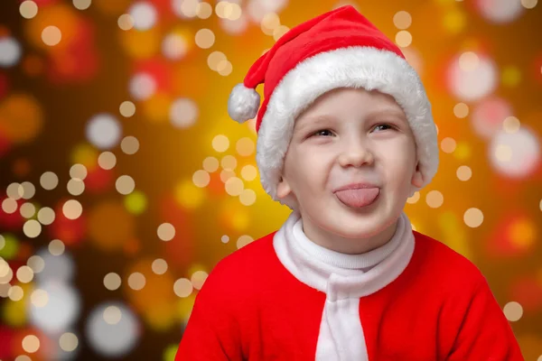 Pequeño Santa se sienta en medio de las luces de Navidad y sonriente muestra la lengua — Foto de Stock