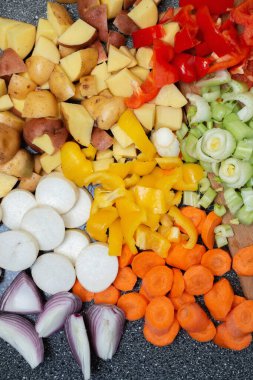 Kesme tahtasındaki sebzeler. Ev mutfağı, yemek pişirme sürecinin yakın çekimi. Taze sebze doğradım. Havuç, kereviz, kırmızı soğan, kırmızı ve sarı biber. Beyaz ahşap masadaki tahta kesme tahtasına yerleştirilmiş, yukarıdan görülüyor. Düz la