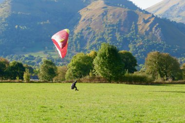 Yere canlı kırmızı ve sarı bir gölgelik üzerinde bir paraglider yaklaşıyor. Sahnede açık mavi gökyüzünün altında yemyeşil tarlalar ve uzak dağlar yer alıyor..