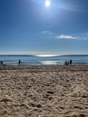 Bir grup insan sahilde oynarken ve dinlenirken diğerleri suyun kenarında yürürken görülüyor. Güneş yukarıdaki mavi gökyüzünde parlıyor..