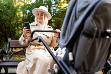 Elinde akıllı bir telefon olan mutlu anne parkta bir bankta oturur ve bebeği bebek arabasında sallar.