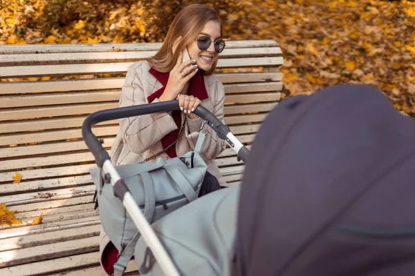 Stylish mom with stroller talking on the phone while sitting on  bench in the park while the child is sleeping