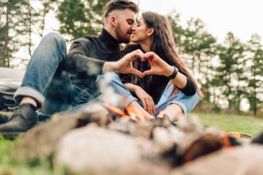 Kamp ateşinin yanında oturan romantik çift öpüşüyor ve kalp şeklinde jest yapıyor.