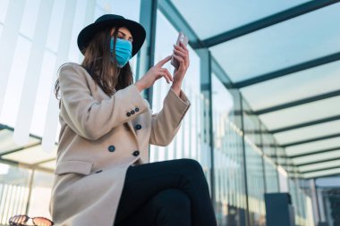 Koruyucu maskeli kız otobüs durağında elinde telefonla oturuyor. Kadın salgın sırasında otobüs durağında oturur ve nakliyeyi bekler.