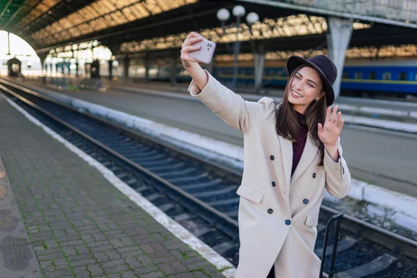 Female tourist talking on video call while standing on the platform of the railway station. Woman waving hand at phone camera