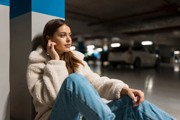 Fashionable girl listening to music in wireless headphones while sitting in the underground parking of shopping mall