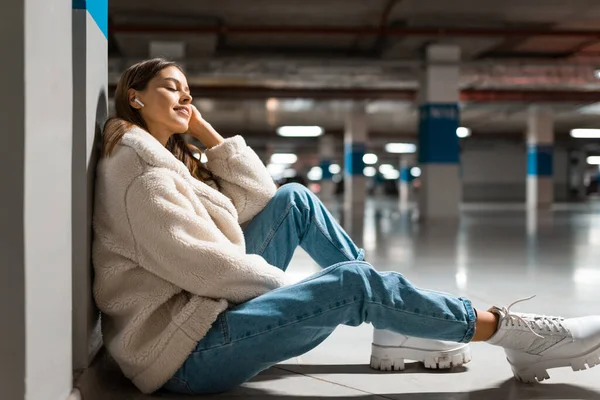 Fashionable girl listening to music in wireless headphones while sitting in the underground parking of shopping mall