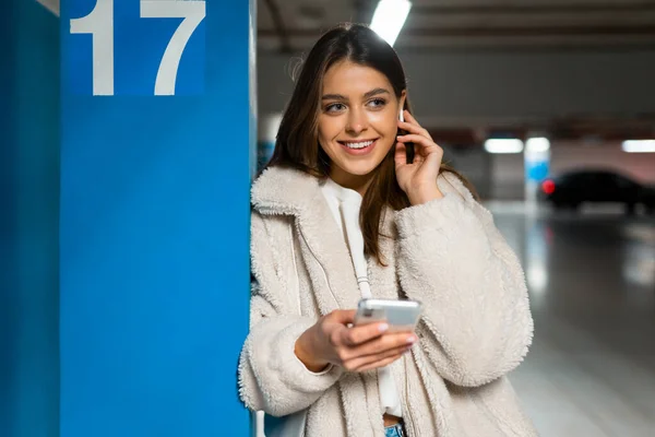 Portrait of smiling girl with phone in hands who correcting the earphone in her ear. Young fashionable woman with phone and headphones at the underground parking