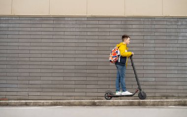 Sırt çantalı modern bir genç, arka planda elektrikli scooter kullanıyor. Çocuk okuldan dönüyor.