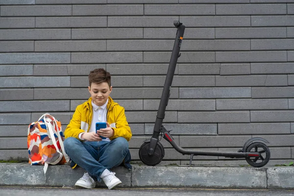 在他的电动车和背包旁边坐着一个拿着手机的青少年 现代男孩在智能手机上聊天 — 图库照片