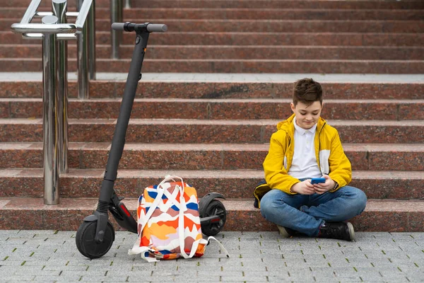 现代青少年坐在他的电动车旁边的楼梯上 用智能手机发短信 — 图库照片