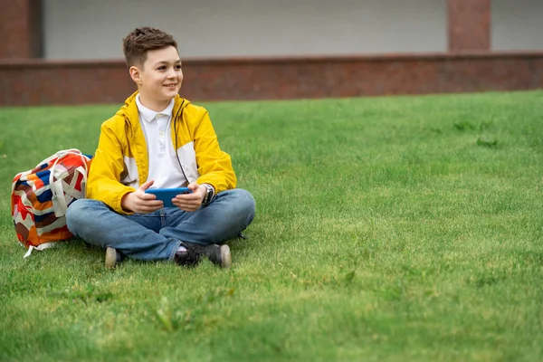 Schoolboy sits on the lawn with  smart phone in his hands. Modern teenager in yellow jacket