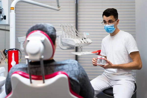 Dental surgeon with dental crown in hands model talks to patient about prosthetics. Man at the dentist\'s appointment