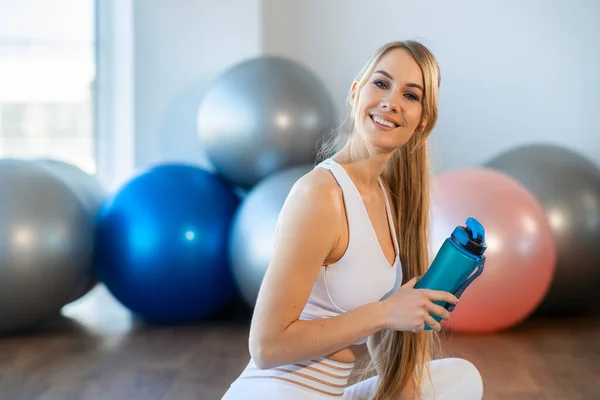 Portrait of beautiful fitness woman sitting on mat with sports bottle in her hands