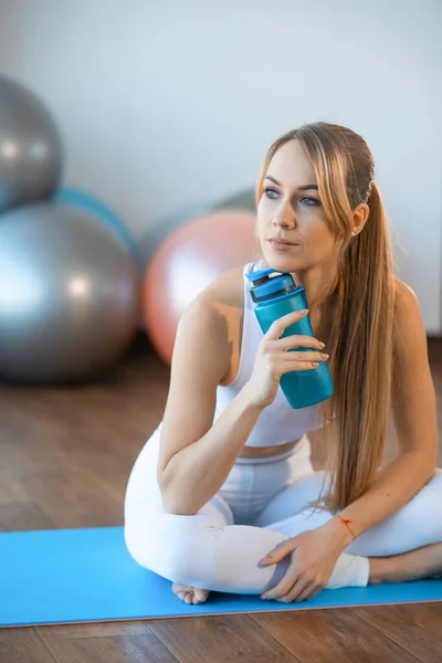 Pensive girl in sportswear sitting on the floor in the gym with sports bottle in her hand