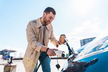 Elinde kahve fincanı olan şık bir adam elektrikli arabanın prizine takar.