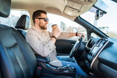 Başarılı bir adam elinde bir fincan kahveyle araba sürüyor.