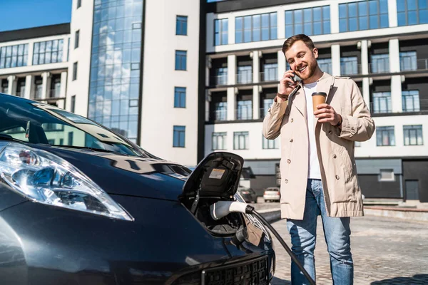 Handsome man talks on the phone and drinks coffee while his car is charging at the station