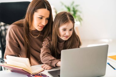 Anne ve kızı laptopta ödev yapıyor. Uzaktaki küçük okul kızı evde öğreniyor.