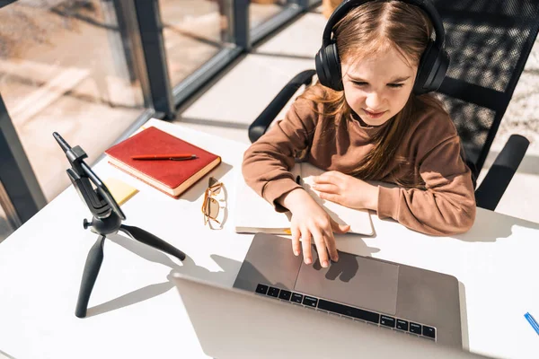 Cute schoolgirl in headphones works at laptop. Little girl study online