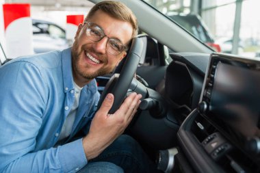 Tatmin olmuş bir adam kafasını yeni arabasının direksiyonuna koydu. Otomobil galerisinde gözlüklü bir erkek.