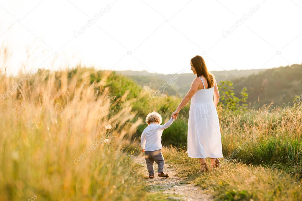 Back view of a beautiful woman in a light summer dress walks with her son on a picturesque nature