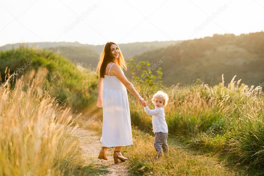Pretty mother in white summer dress walks with her beautiful son along the path of a picturesque field