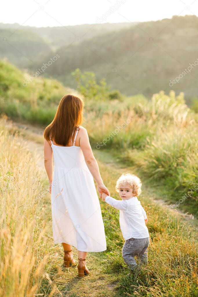 Elegant woman in white dress leads her son by the hand while walking in nature