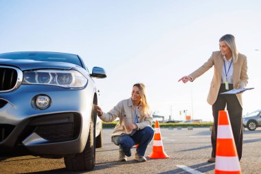 Sürücü öğrenci araba lastiklerini kontrol ederken eğitmen ders sırasında ilerlemeyi kaydediyor