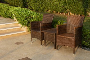 Parkta dinlenmek için rattan mobilyalar. Yeşil çalıların arkasındaki sandalye ve masa.