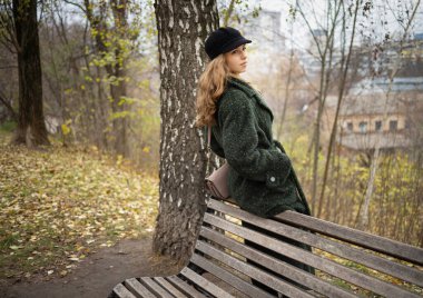 Uzun kıvırcık saçlı, yeşil ceketli, siyah şapkalı şık bir kız. Şehir parkında bankın yanında.