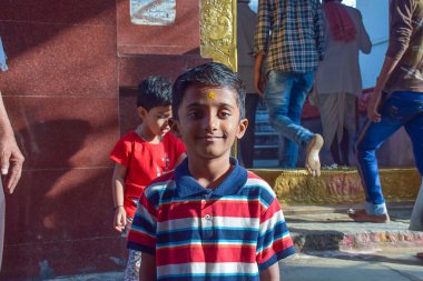 Alnında karalahana tişörtü ve sandal ağacı tilak giymiş, parlak güneşli bir günde şakak merdivenlerinde gülümseyen 5-8 yaşlarındaki Hintli bir çocuğun resmi..