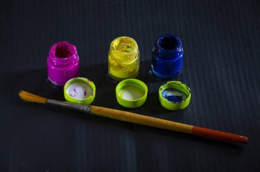 Pembe, mavi, sarı suluboya cam şişenin resmi. Kapakları açık ve siyah arka plandaki şişelerin önünde duran ahşap boya fırçası var..