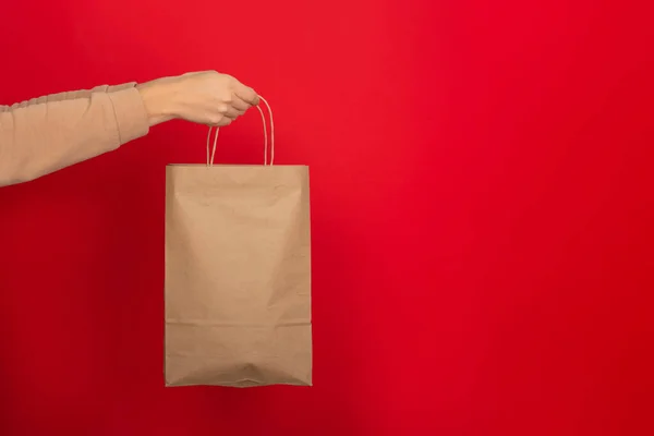 女性の手は赤い背景に茶色のクラフト紙で作られた大きなギフトバッグを保持します ギフト プレゼント またはEショッピングのコンセプト コピースペース付きバナー — ストック写真
