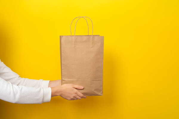 Female hands hold large gift bag made of brown craft paper on yellow background. Gift, present or e-shopping concept. Banner with copy space