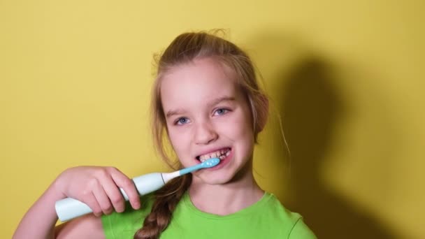 ティーンエイジャーの女の子は黄色の背景に彼女の歯を磨く 緑のTシャツの子供の女の子は電動歯ブラシを使用しています — ストック動画