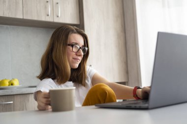 Modern mutfakta dizüstü bilgisayarda çalışan bir kadın. Merkez ofis konsepti. Kız, dizüstü bilgisayar, modern çatı katında kahve fincanı olan masa..