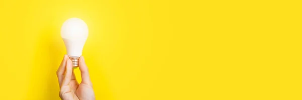 Hand holding a LED light bulb on bright yellow background. Using economical and environmentally friendly light bulb concept. Idea concept. Energy saving lamp in hand. Long Banner with copy space