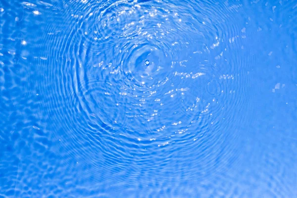 Surface of blue transparent swimming pool water texture with circles on the water. Trendy abstract nature background. Water waves in sun light reflections.