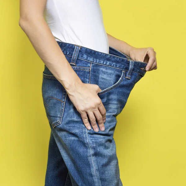 Das Konzept der Ernährung, richtige Ernährung, Gewichtsverlust. Schlanke Frau mit lockeren Jeans und ihrem Abnehmen. Frau weißes T-Shirt und Oversize-Jeans auf gelbem Hintergrund — Stockfoto