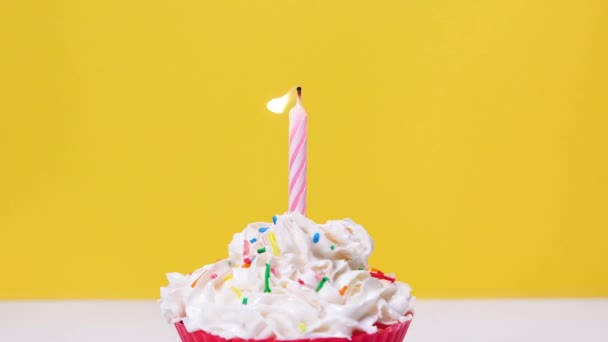 Vista de cerca en sabroso capcake con crema batida y una vela de pastel en llamas aislado sobre fondo amarillo. Soplando una vela de pastel. Full HD cámara lenta feliz cumpleaños o aniversario de vídeo — Vídeo de stock