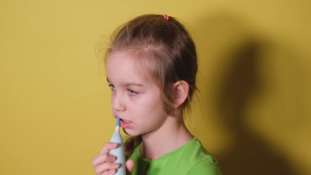 Nastolatka myje zęby na jasnożółtym tle. Dziecko dziewczynka w zielonej koszulce używa elektrycznej szczoteczki do zębów do mycia zębów — Wideo stockowe