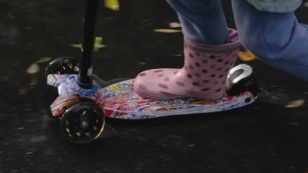 Ein kleines Mädchen in Gummistiefeln fährt auf einem Motorroller über den nassen Asphalt, der mit Pfützen und herabgefallenem Laub bedeckt ist. Full HD Zeitlupe Nahaufnahme Video. — Stockvideo