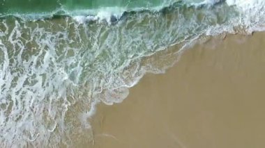 Atlantik Okyanusu 'ndan kumlu bir sahile vuran dalgaların hava aracı videosu. Turkuaz suyun, beyaz deniz köpüğünün ve altın kumun güzel üst manzarası.