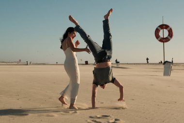 Bir adam kumsalda amuda kalkarken bir kadın izliyor ve gülüyor, neşeli ve kaygısız romantik sahneler..