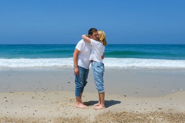 Kumsalda çıplak ayakla duran çift, açık gökyüzü ve günlük kıyafetleriyle dalgaların yanında sarılıyorlar..