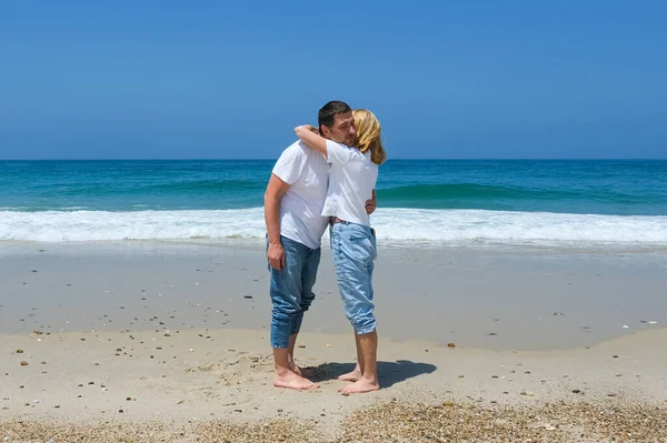 Kumsalda çıplak ayakla duran çift, açık gökyüzü ve günlük kıyafetleriyle dalgaların yanında sarılıyorlar..